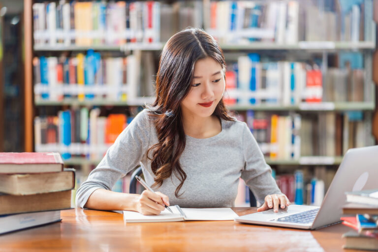 Asian young Student in casual suit doing homework and using technology laptop in library of university or colleage with various book and stationary over the book shelf background, Back to school